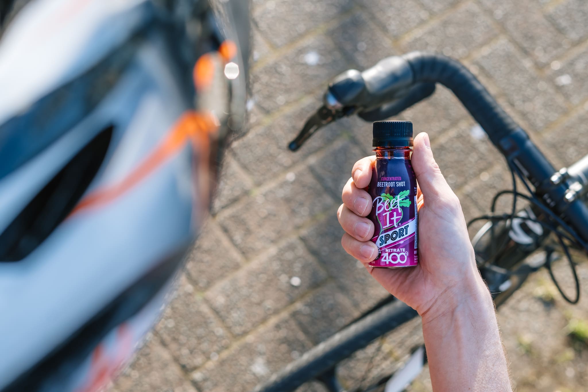 Cyclist holding bottle of Beet It Sport Nitrate 400 next to a bicycle on a paved surface
